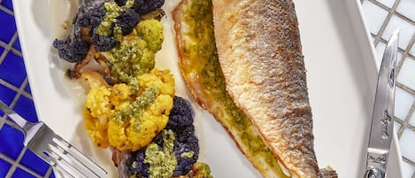 En tallrik med grillad fisk, rostad blomkål och en citronskiva, placerad på ett bord med kakelplattor.