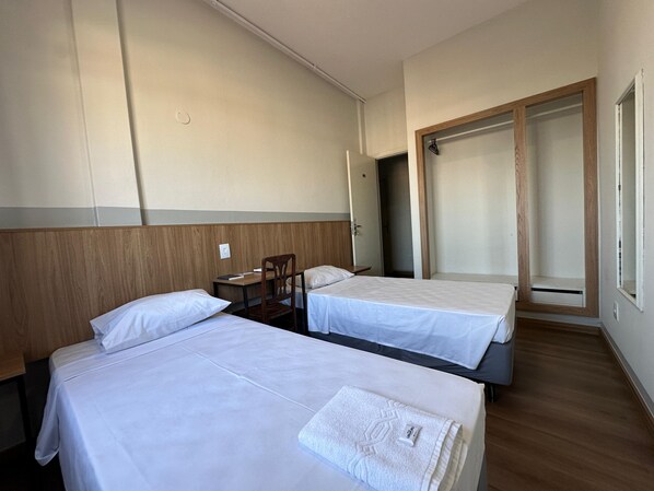 A hotel room with two single beds, a wooden headboard, a desk with a chair, and a wardrobe.