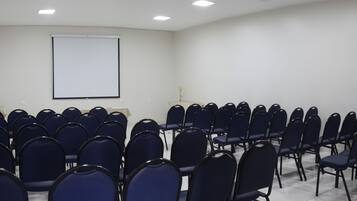 Sala de conferência com fileiras de cadeiras azuis, uma tela de projeção branca e duas plantas.