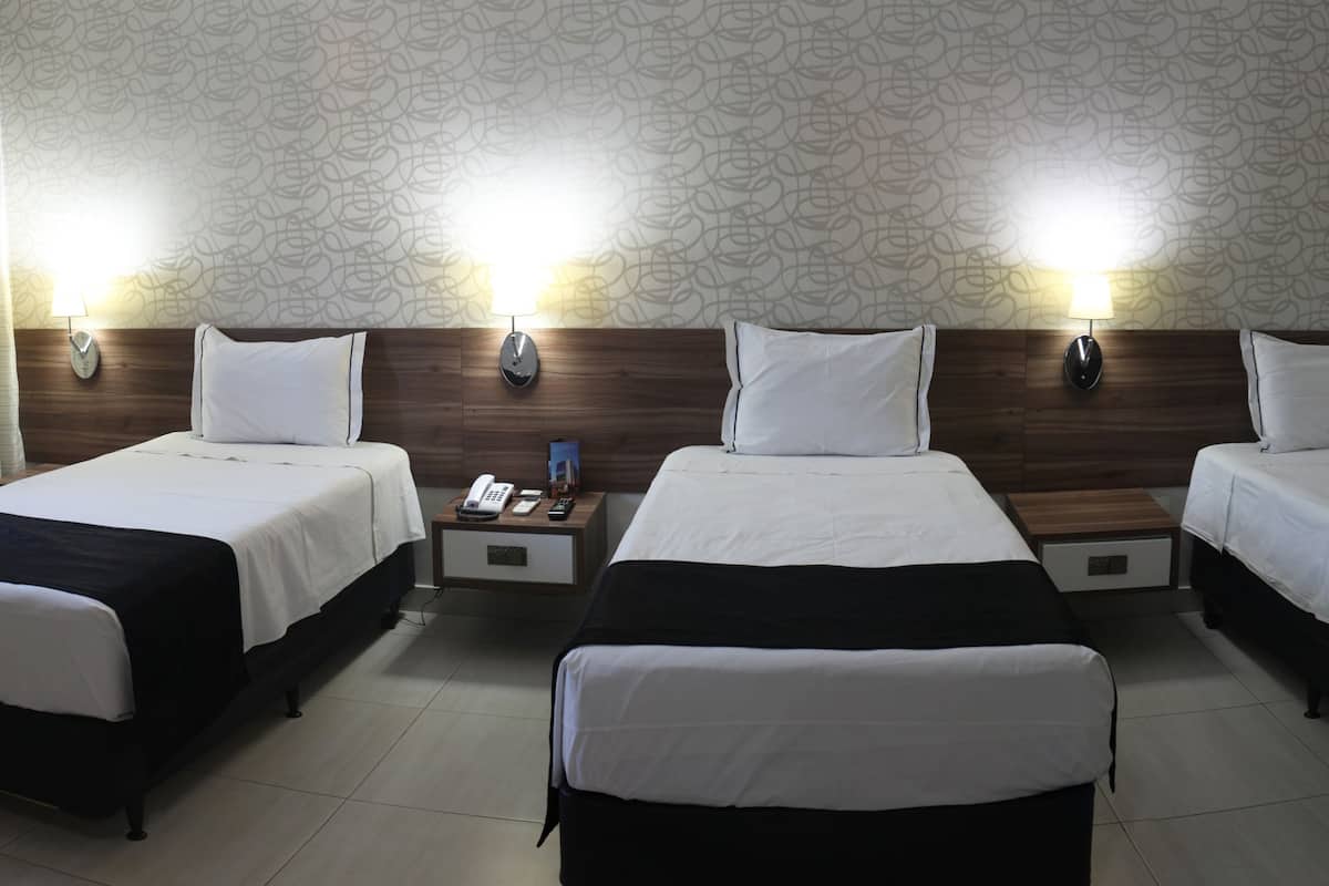 Ett hotellrum med tre separata sängar, var och en med en sänggavel i trä och sänglampor. Mellan sängarna står ett litet bord med en telefon och en fjärrkontroll.