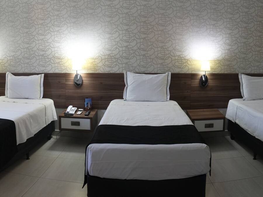 A hotel room with three single beds, each with a wooden headboard and bedside lamps. There is a nightstand between the beds with a phone and a remote control.