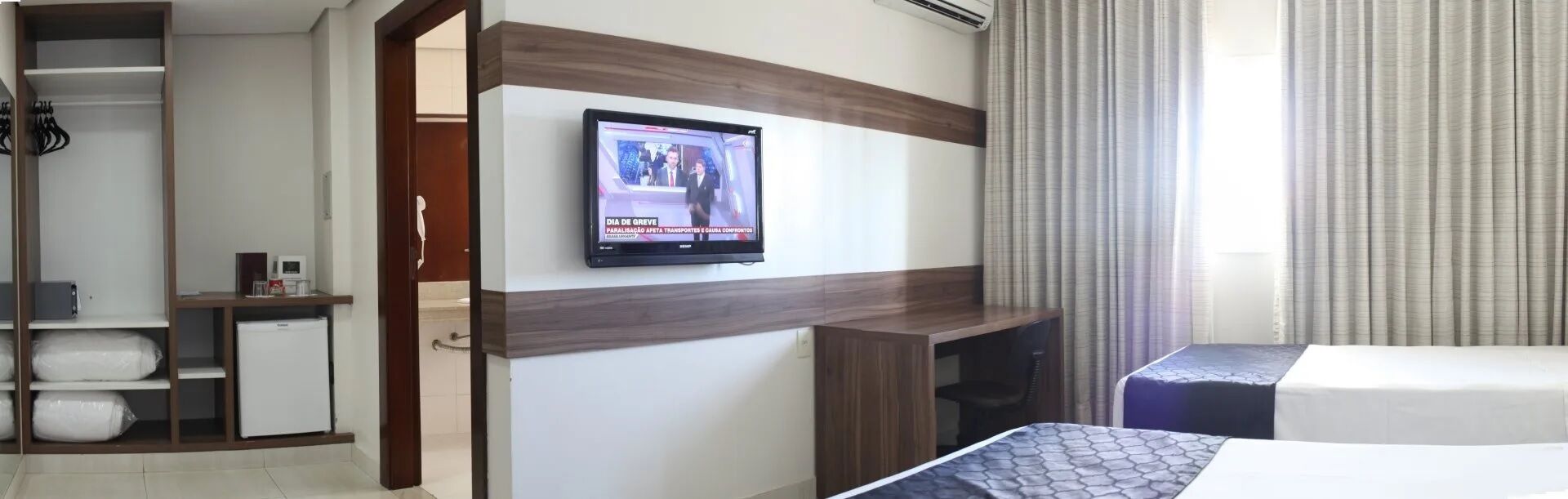 A hotel room with a television mounted on the wall, a bed with a patterned bedspread, a desk with a computer, and a wardrobe.