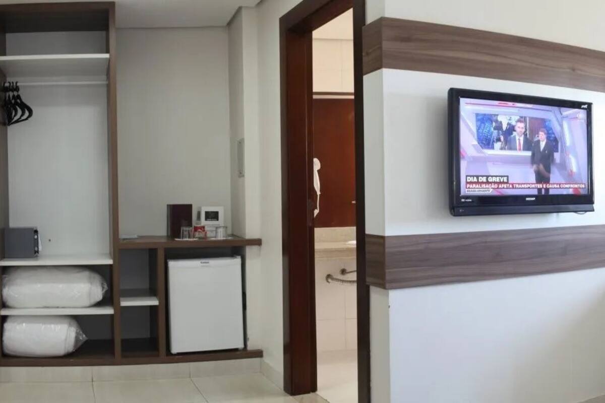 Ett hotellrum med en tv som är monterad på väggen, en säng med ett mönstrat överkast, ett skrivbord med en dator och en garderob.