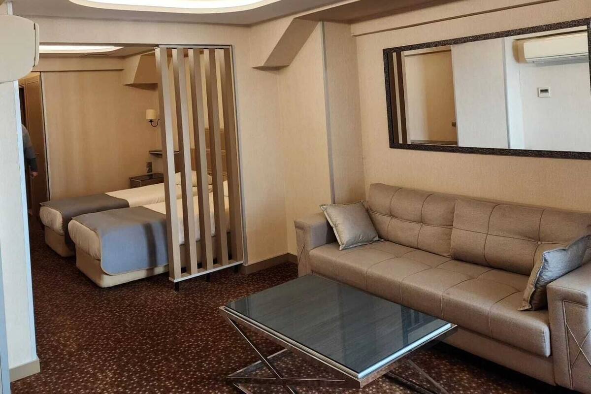 A modern hotel room with a sofa, a glass coffee table, a mirror, and a ceiling light fixture.