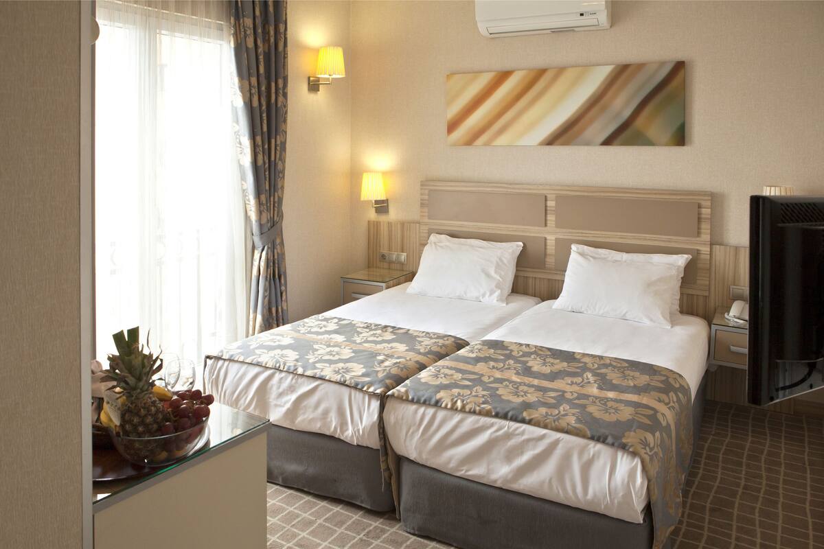 A hotel room with two beds, a flat-screen TV, a bedside table with a lamp, and a fruit bowl on the windowsill.