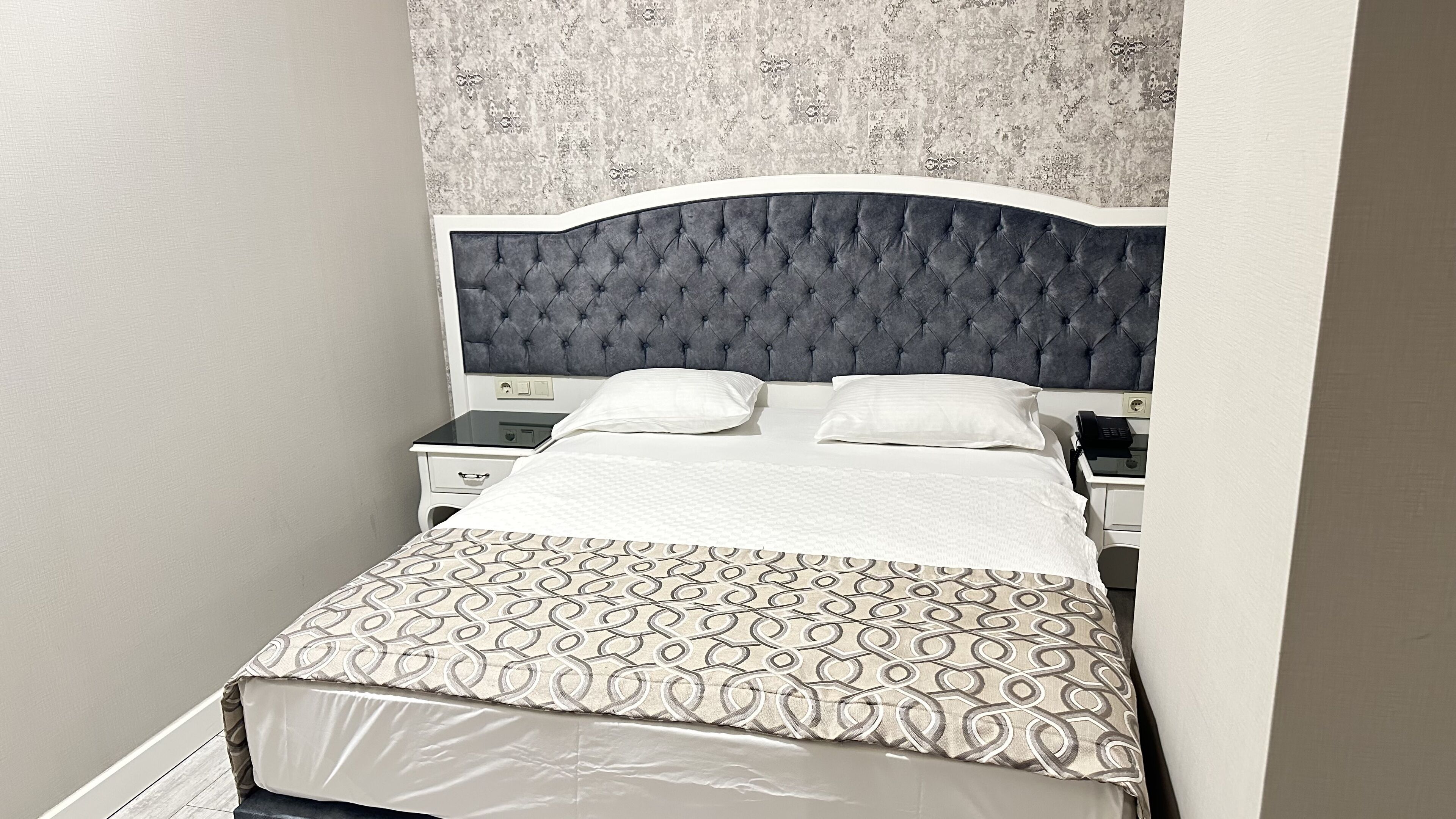 A neatly made bed with a patterned bedspread, a tufted headboard, and bedside tables with lamps and a phone.