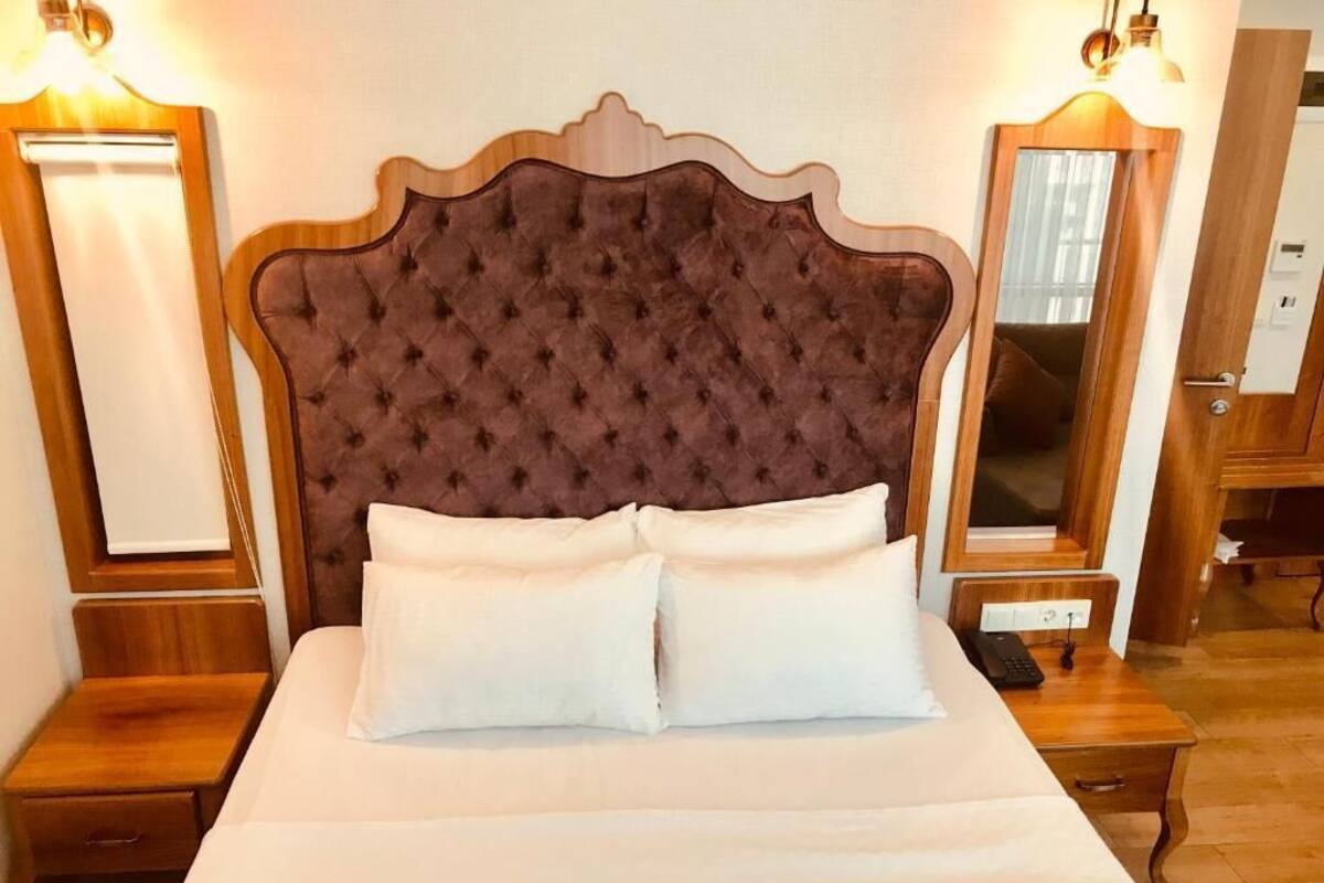 Een opgemaakt bed met een gestoffeerd hoofdbord, twee nachtkastjes en een spiegel aan weerszijden.