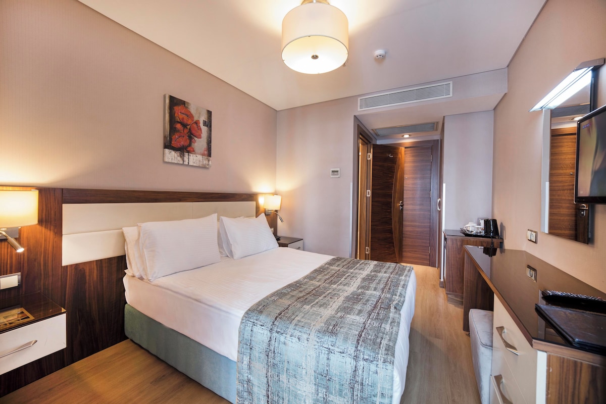 Habitación de hotel con cama, mesitas de noche, escritorio, silla, televisor y puerta que da a otra habitación.