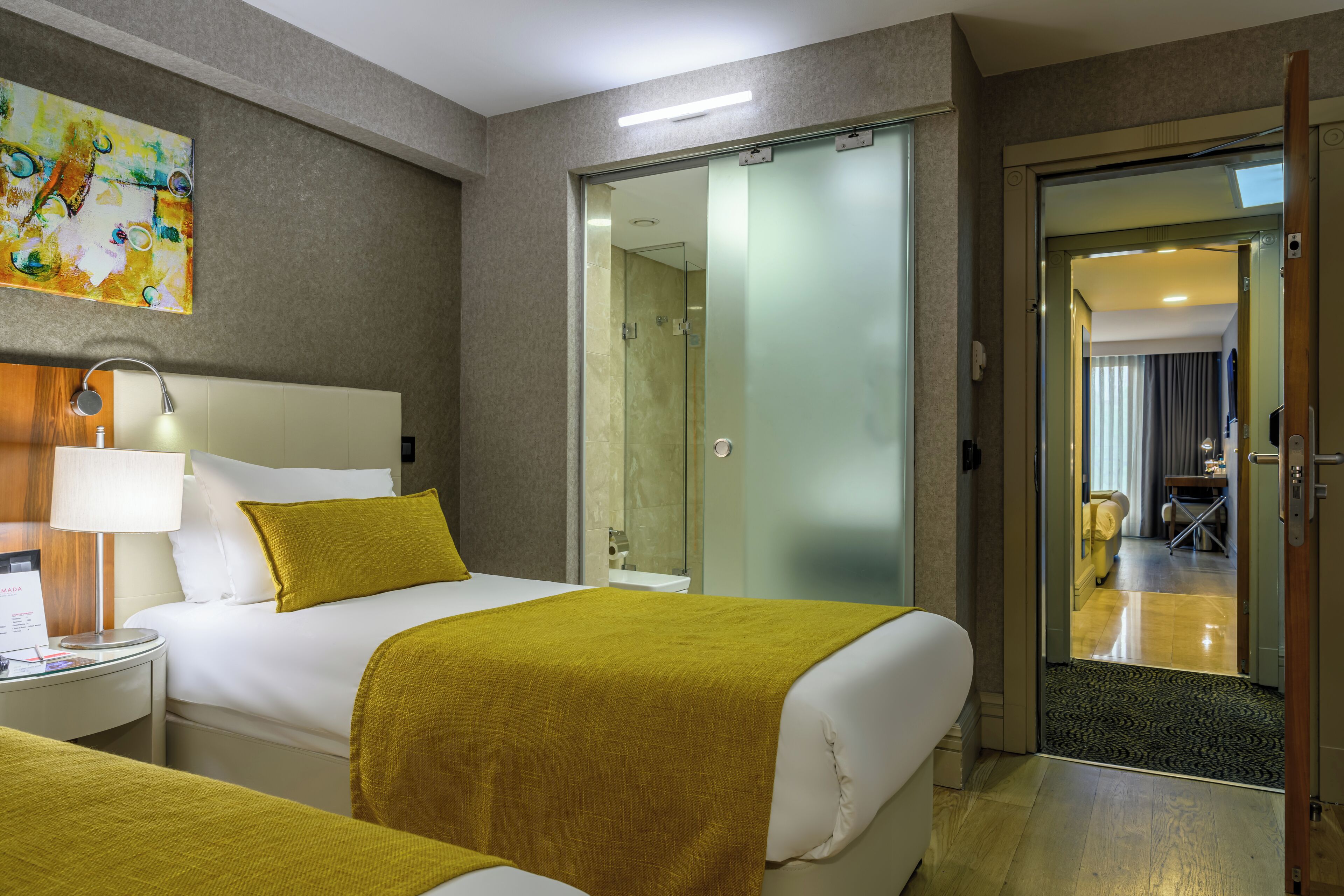 A hotel room with a bed, a bedside table with a lamp, a glass shower enclosure, and a door leading to a balcony.
