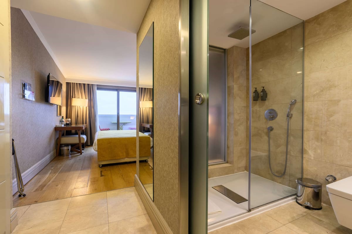 A modern hotel room with a glass shower enclosure, a bed, a desk with a lamp, and a view of the sea.