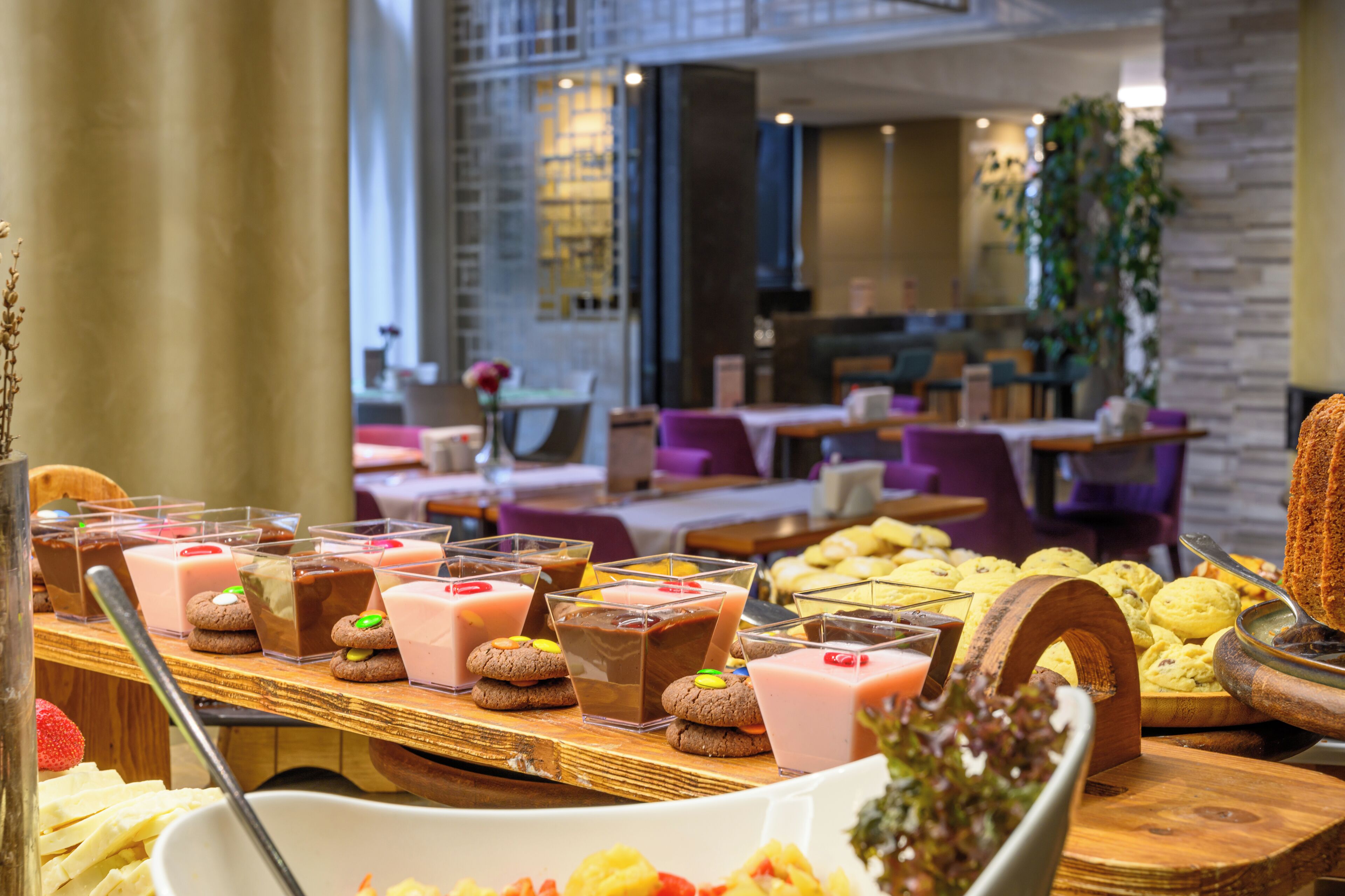 A buffet with various desserts and drinks, including cookies and chocolate milkshakes, in a modern restaurant setting with a stone wall and indoor plants.