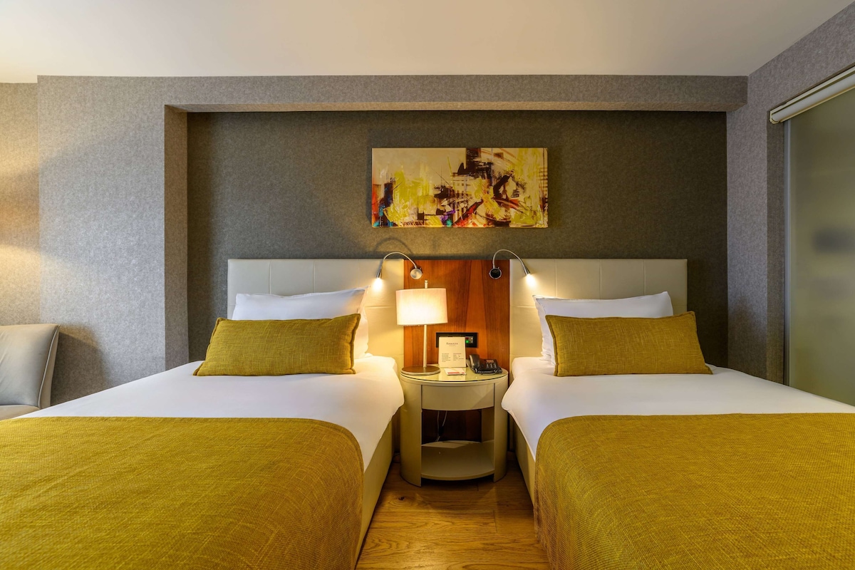 A hotel room with two beds, a nightstand with a lamp, and a painting on the wall.