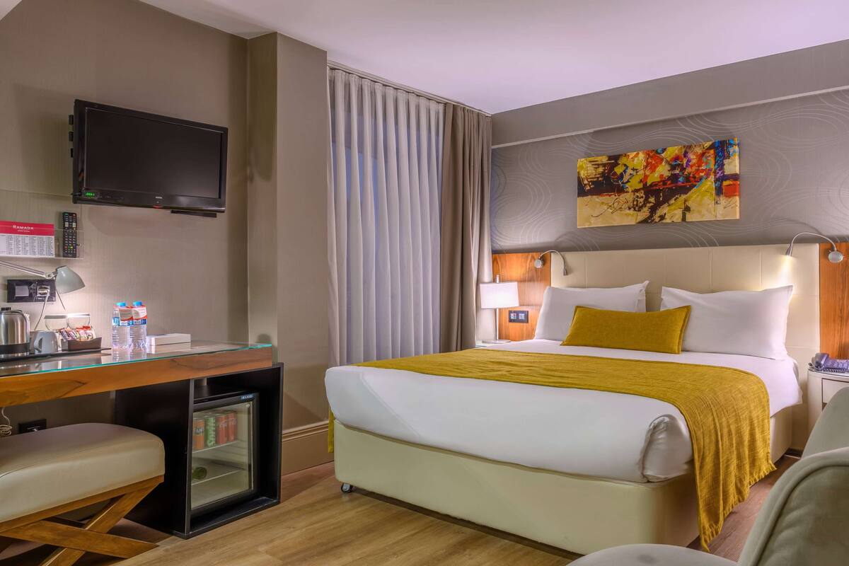 A hotel room with a bed, a desk with a computer, a mini-fridge, a TV, and a painting on the wall.
