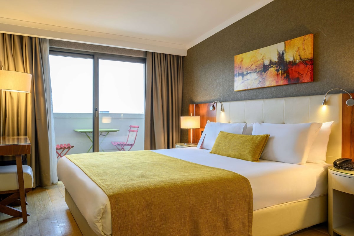 A hotel room with a large bed, a desk, a chair, a balcony with a table, and a painting on the wall.