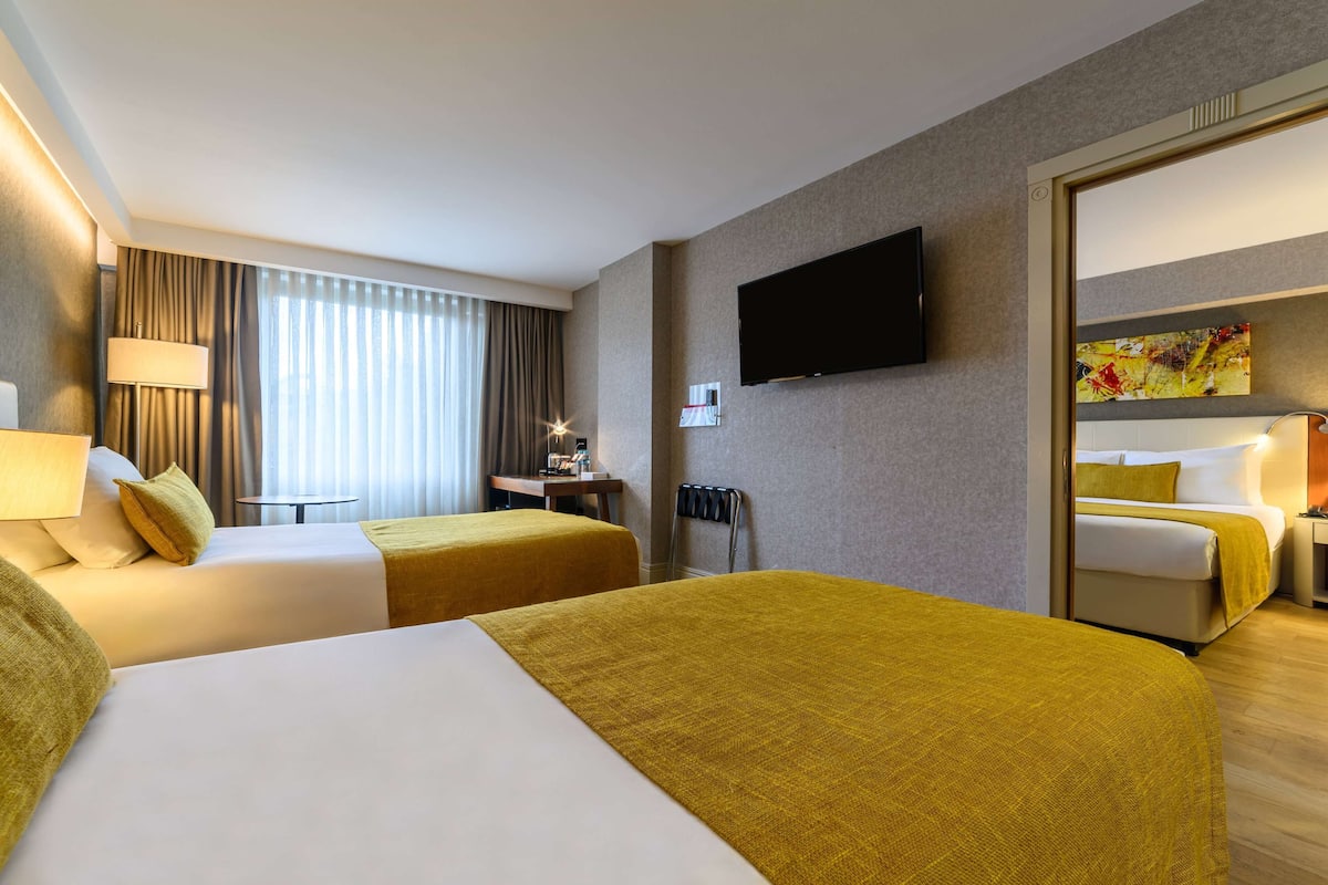 A hotel room with two beds, a TV mounted on the wall, a desk with a chair, and a window with curtains.