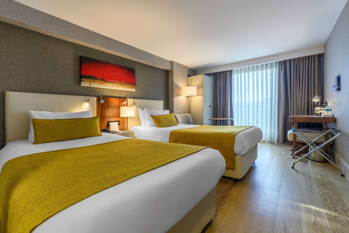 A hotel room with two beds, a desk, a chair, a lamp, and a large window with curtains.