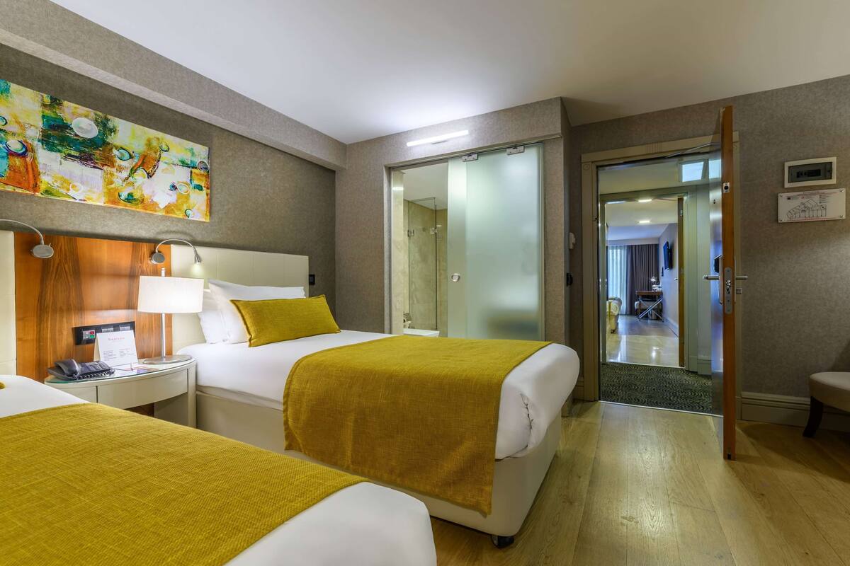 A hotel room with two beds, a wooden headboard, a bedside table with a lamp, and a painting on the wall.