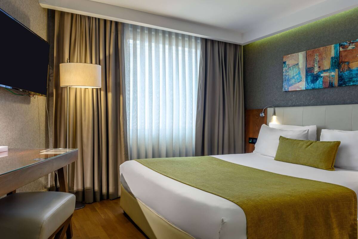 A hotel room with a bed, a desk, a lamp, and a painting on the wall.