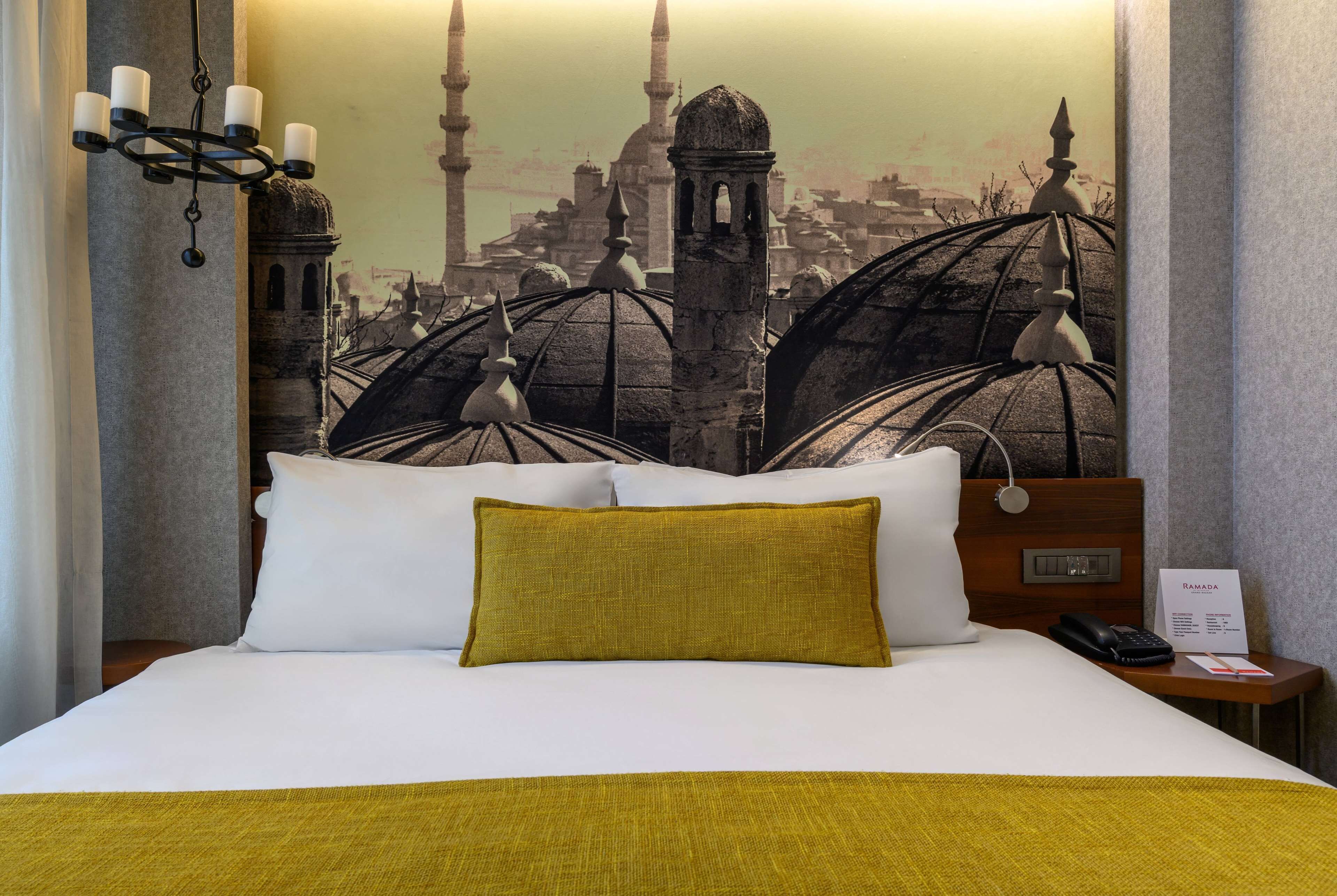 A hotel room with a bed, a nightstand, a lamp, and a wall tapestry depicting a cityscape.