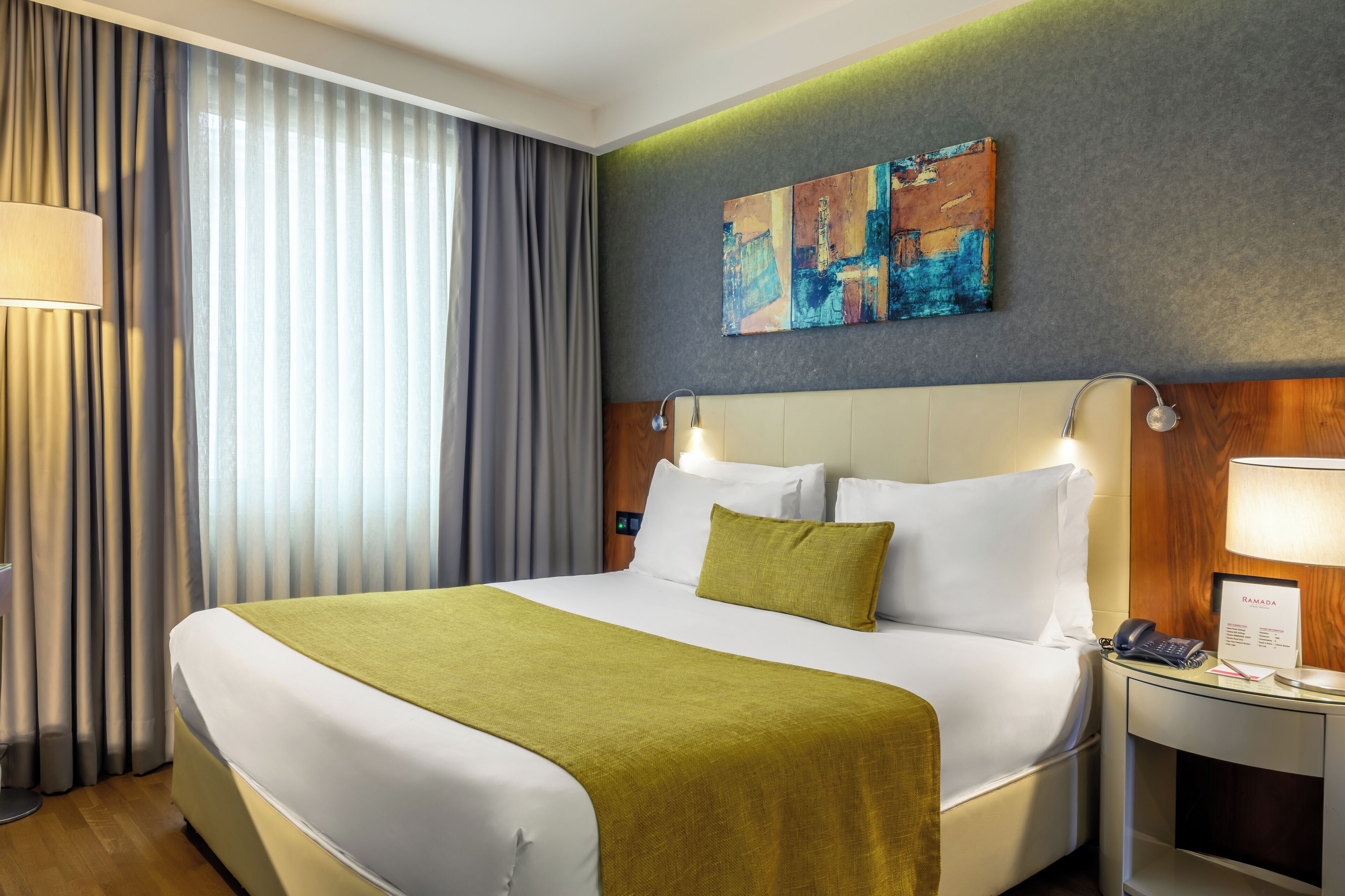 A hotel room with a large bed, a bedside table with a lamp, a wall-mounted artwork, and a window with curtains.