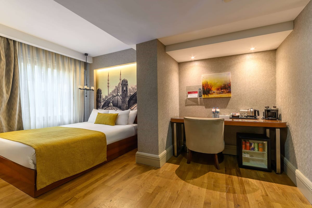 A hotel room with a bed, a desk, a chair, a refrigerator, and a painting on the wall.