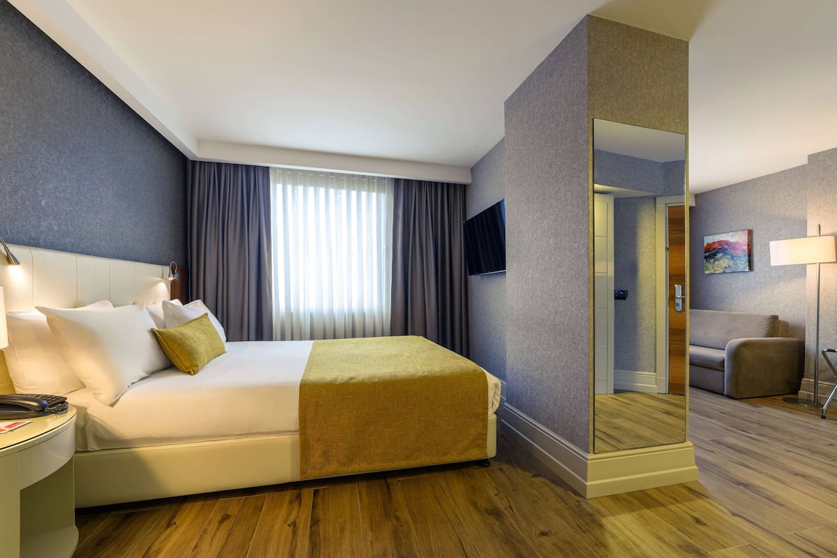 A hotel room with a bed, a desk with a lamp, a TV mounted on the wall, and a mirror.