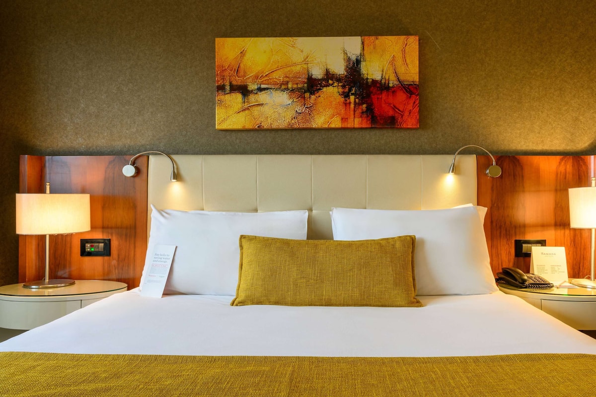 A hotel room with a large bed, two bedside tables, a nightstand, a wall-mounted painting, and a headboard with integrated lighting.