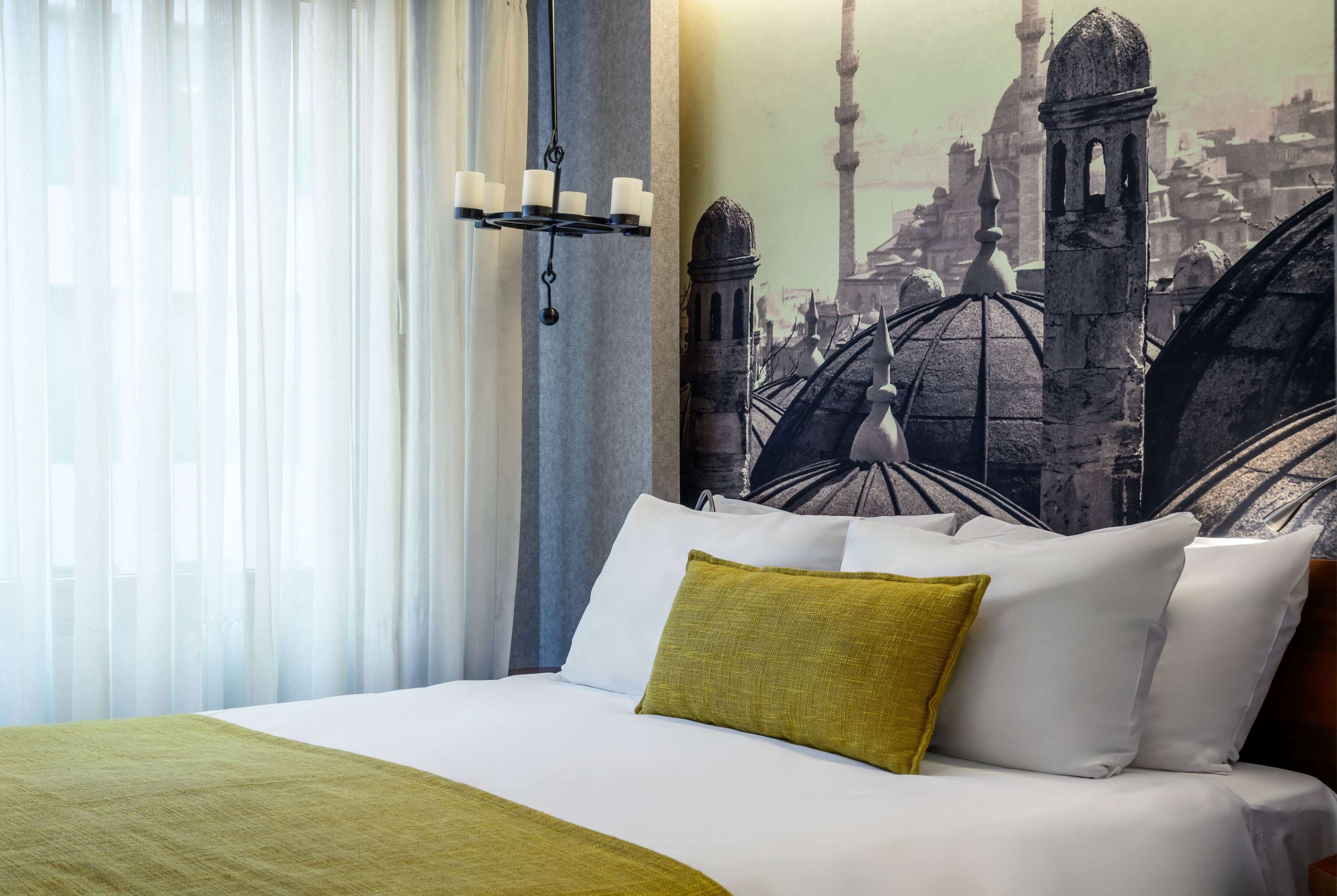 Ein Hotelzimmer mit einem Bett, einer grünen Tagesdecke, weißen Kissen, einer Kristallleuchtre und einem großen Wandgemälde einer Stadtansicht.