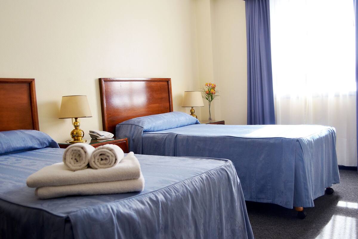 A hotel room with two beds, each with a blue bedspread and matching pillows. There are two bedside tables with lamps and a vase of flowers. The room has a wooden headboard and a window with blue curtains.