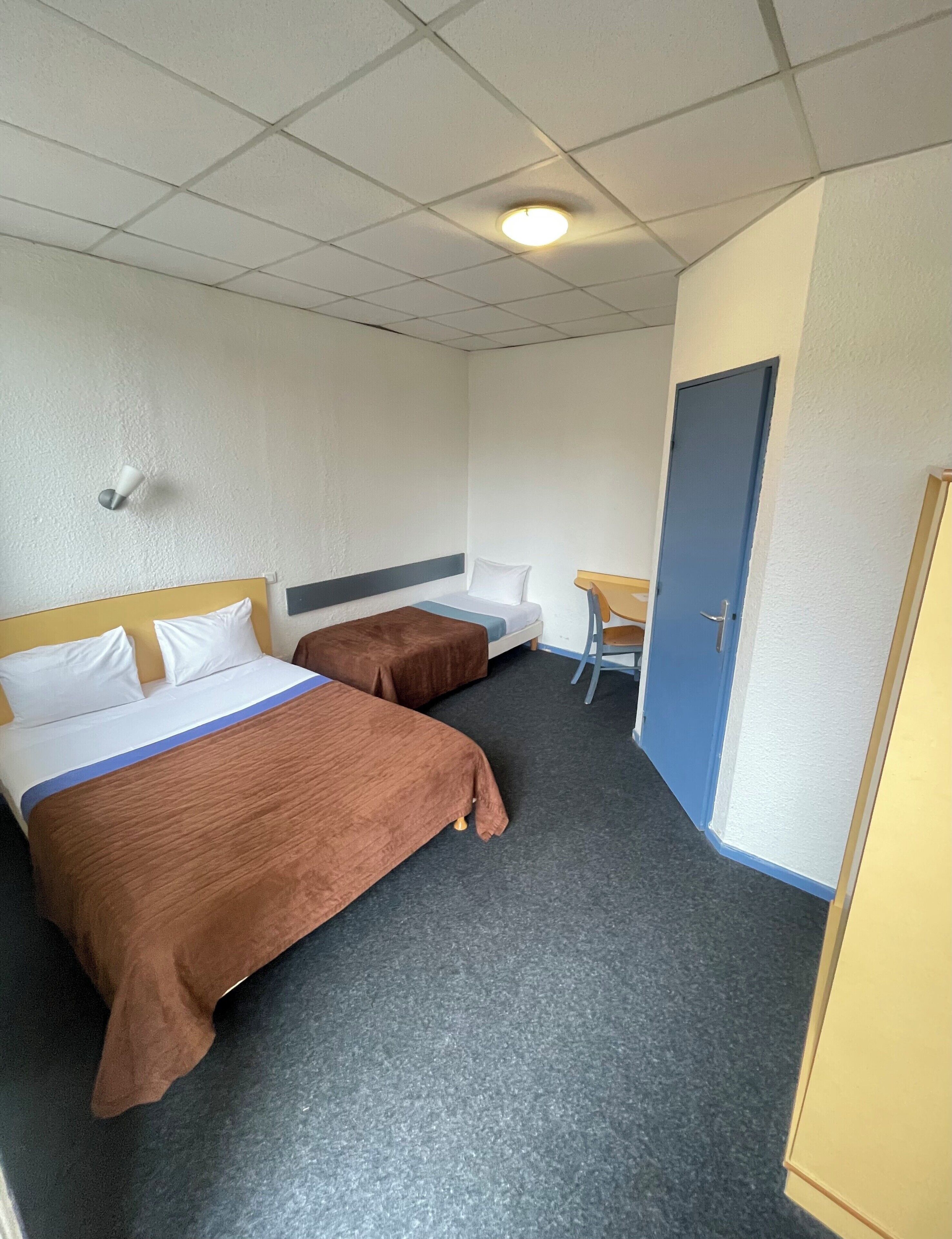 A hotel room with two beds, a desk, a chair, a door, and a ceiling light.