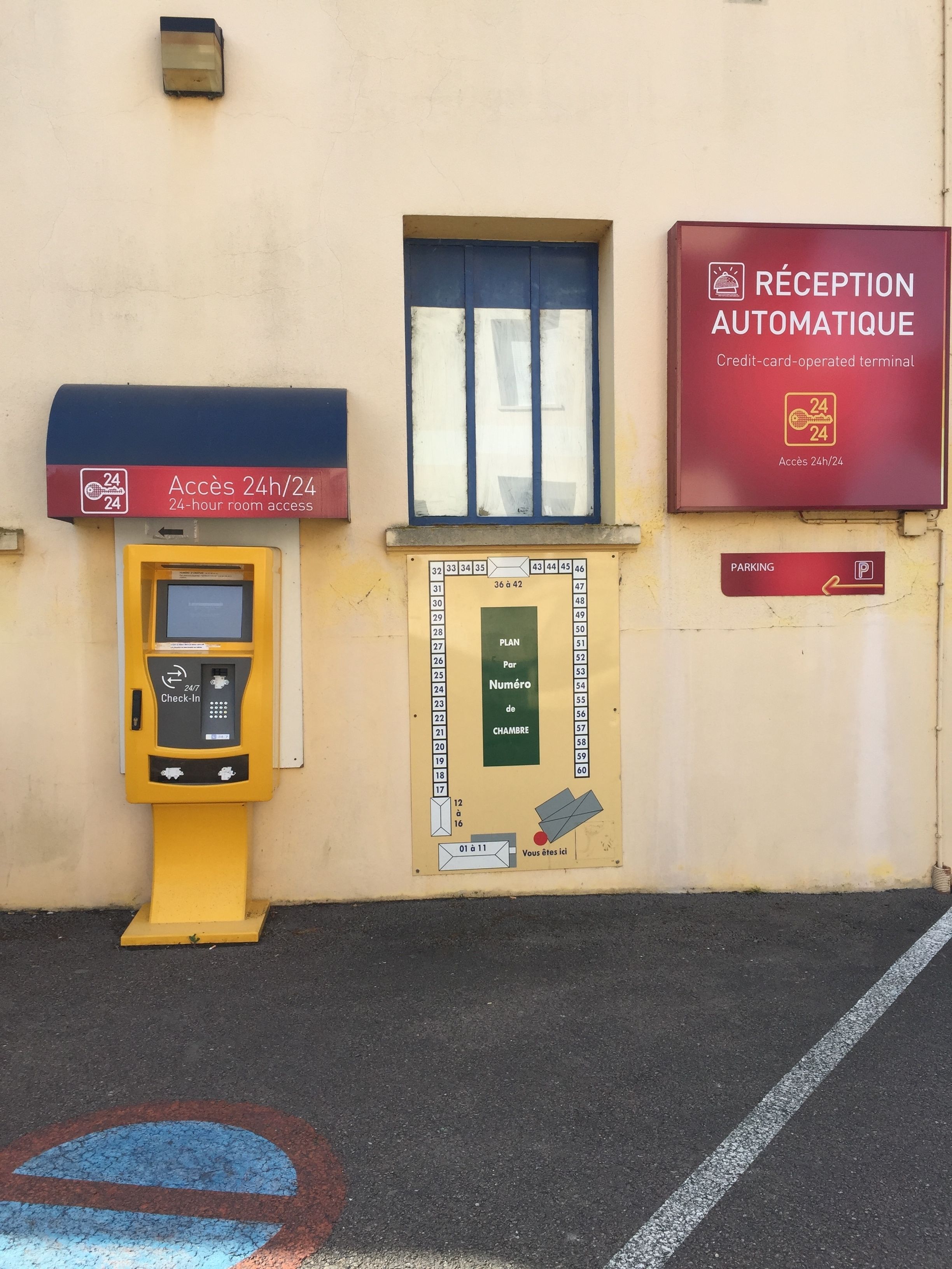 A hotel reception area with a 24-hour access sign, a check-in kiosk, a room number board, and a parking sign.
