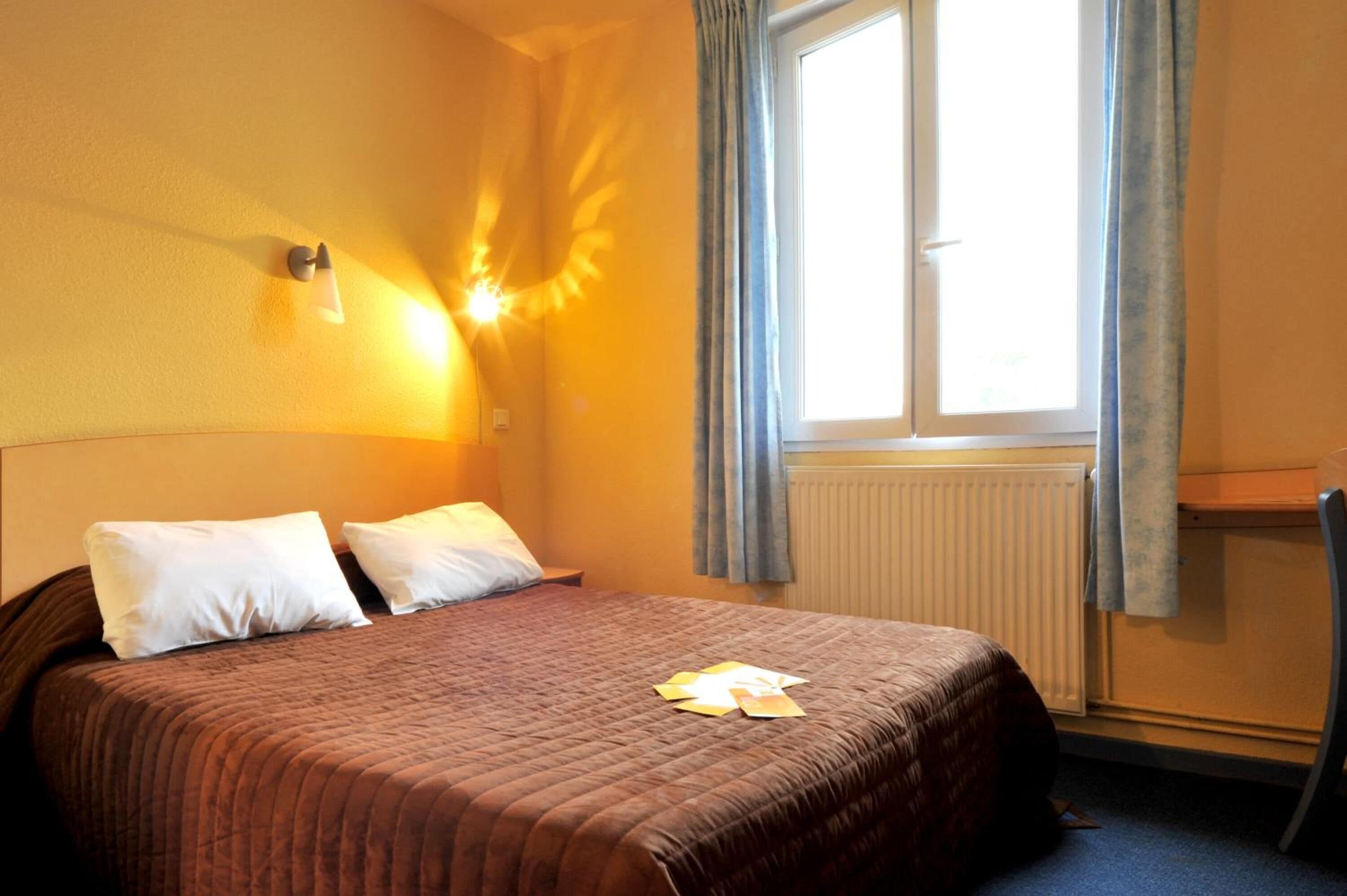 A hotel room with a bed, a nightstand, a window with curtains, and a wall-mounted light fixture.