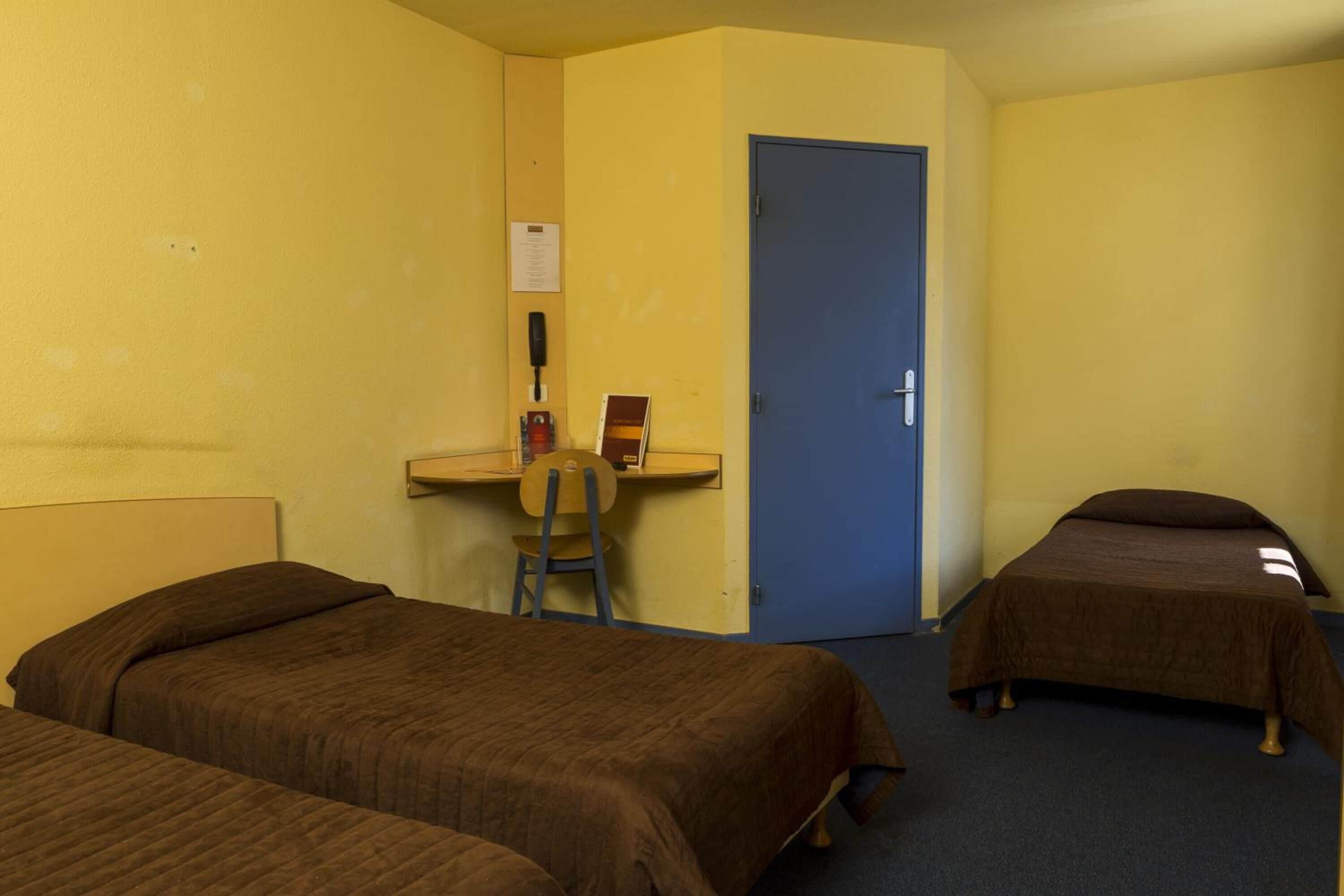 Une chambre avec deux lits simples, un bureau avec une chaise et une porte bleue.