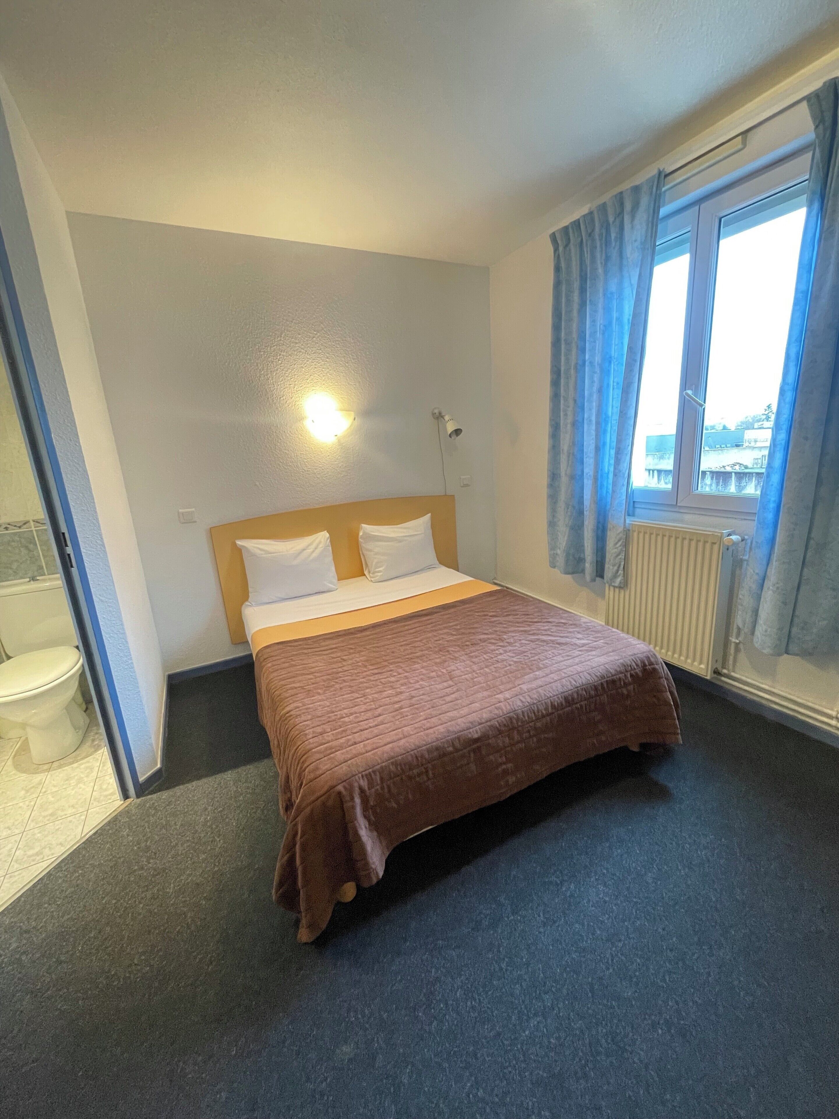 Habitació d'hotel amb llit, finestra amb cortines, radiador i llum penjada a la paret.