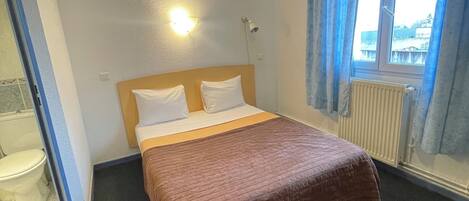 A hotel room with a bed, a window with curtains, a radiator, and a wall-mounted light.