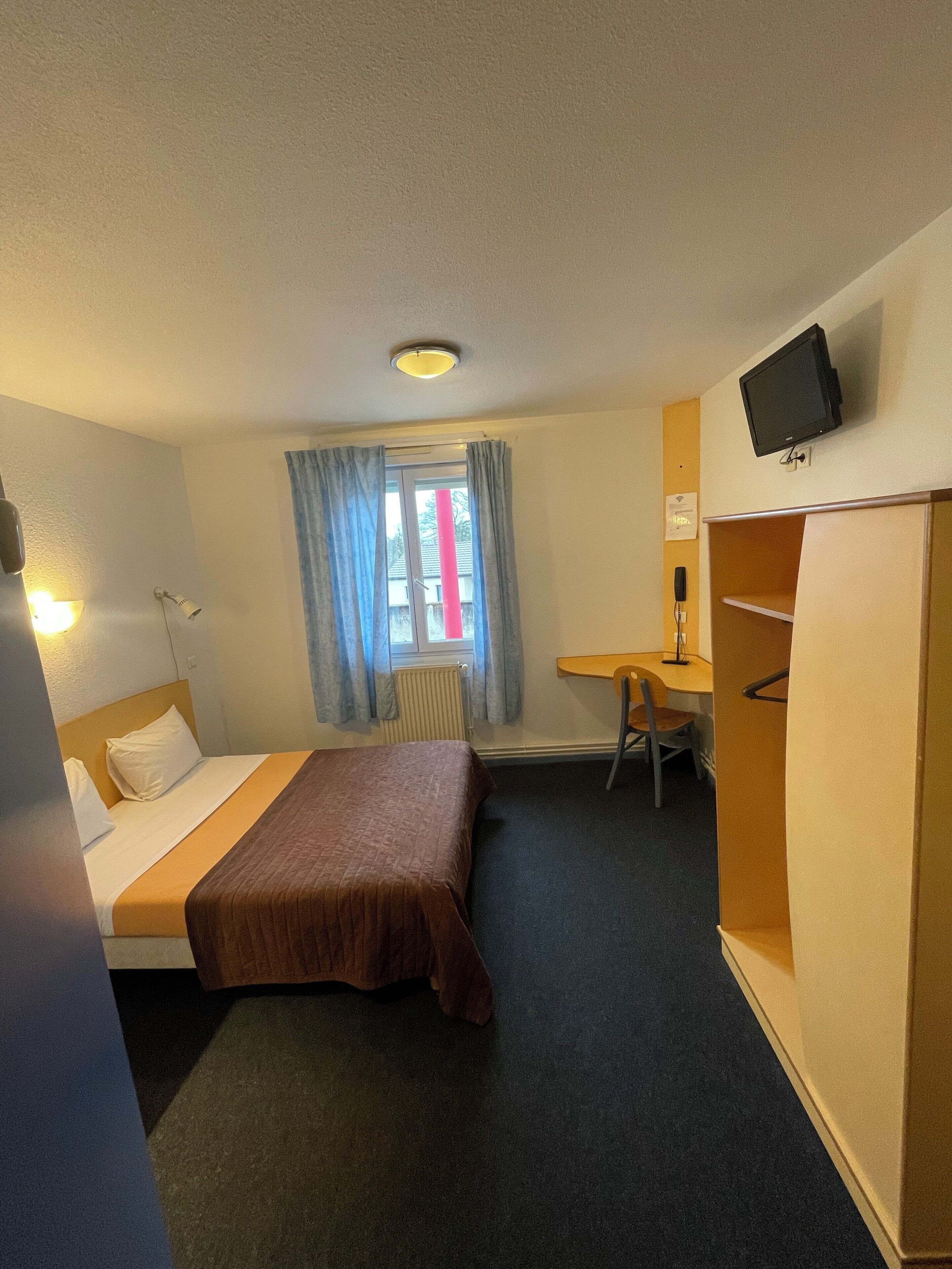 A hotel room with a bed, a desk, a chair, a television, and a wardrobe.