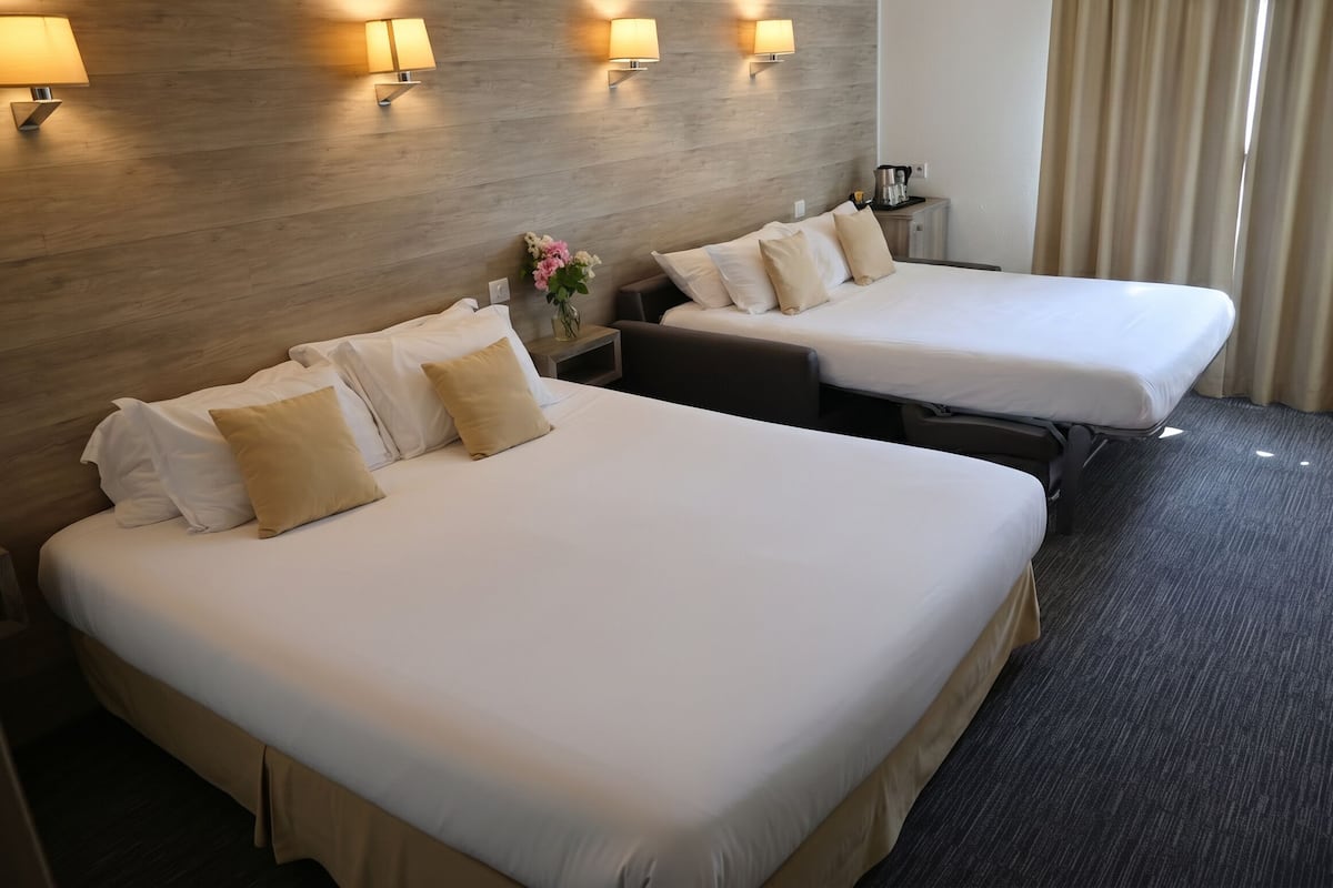 A hotel room with two beds, a wooden headboard, wall-mounted lamps, a nightstand, and a vase with flowers.