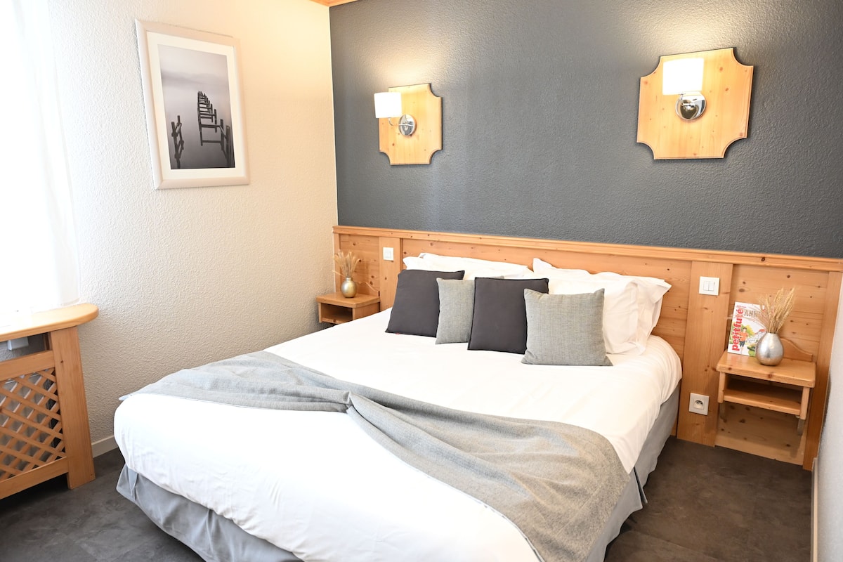 A neatly made bed with a grey and white duvet, wooden headboard, two bedside tables with lamps, a framed picture on the wall, and a small wooden shelf with a vase.
