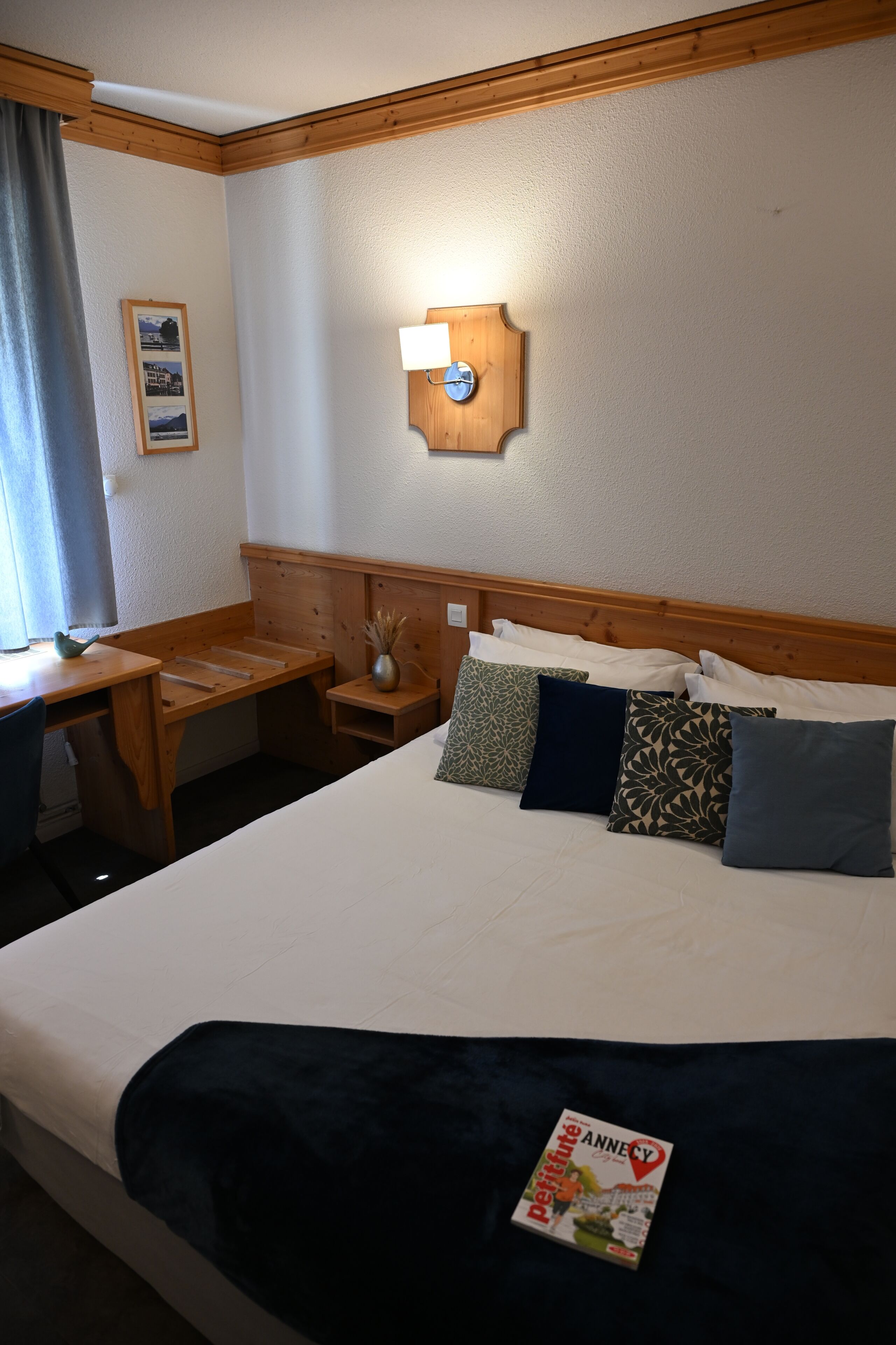 A hotel room with a bed, a desk, a chair, a nightstand, a lamp, a clock, and a framed picture on the wall.