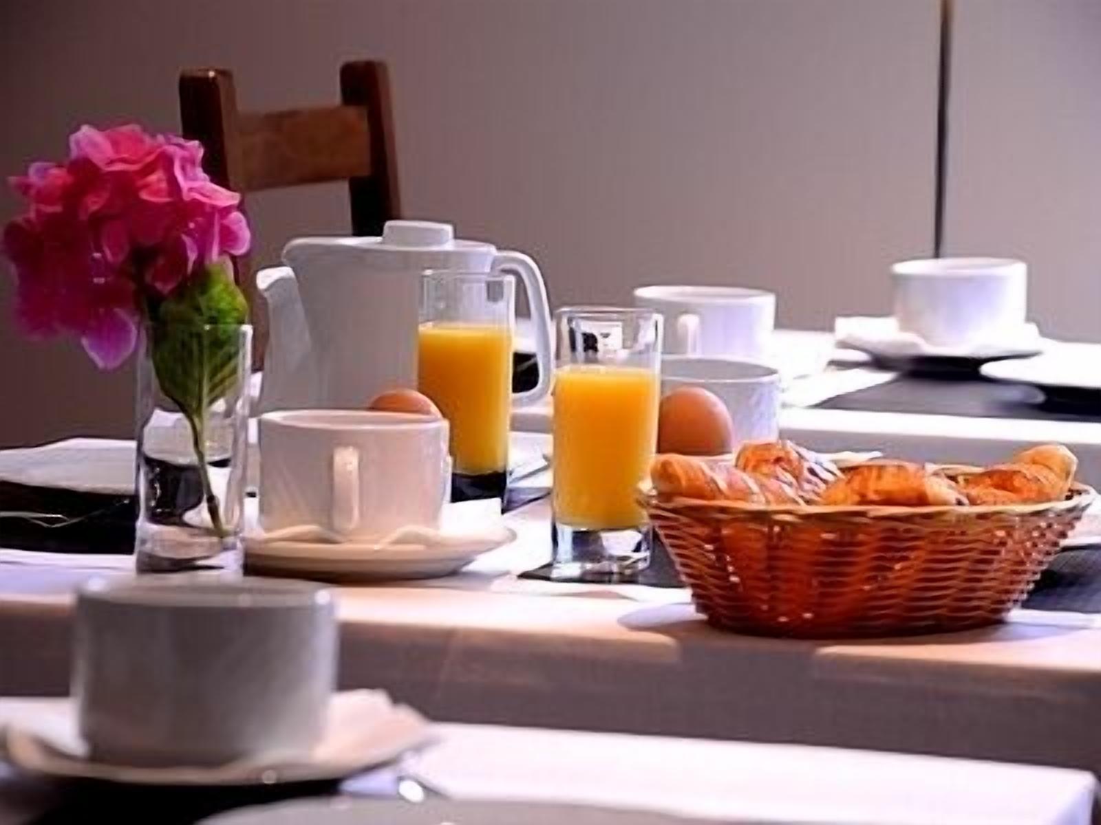 A breakfast setup with a pitcher, cups, a glass of orange juice, a basket of croissants, and a vase with flowers.
