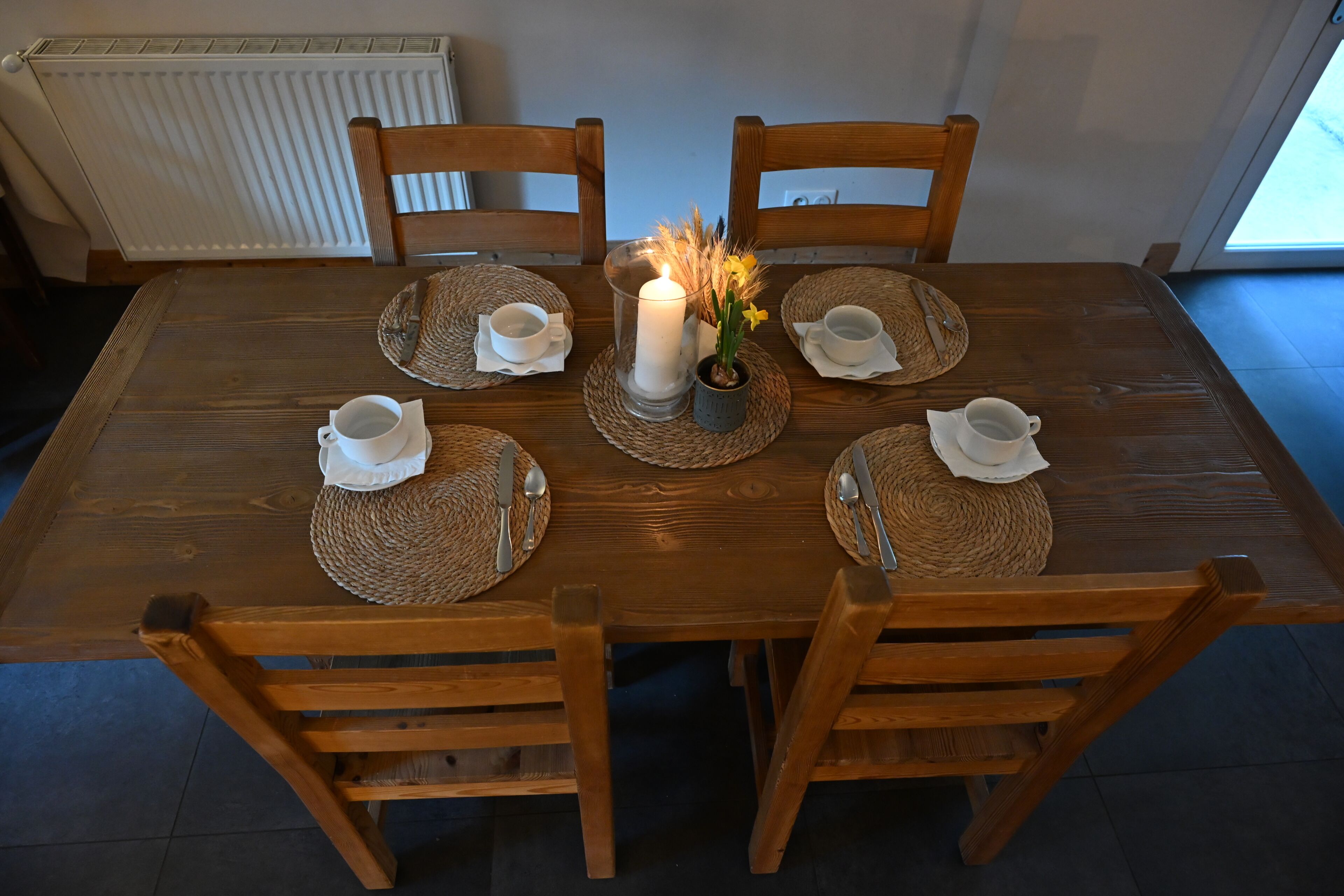Une table à manger en bois, avec quatre chaises, une bougie allumée et un petit vase contenant des fleurs jaunes.