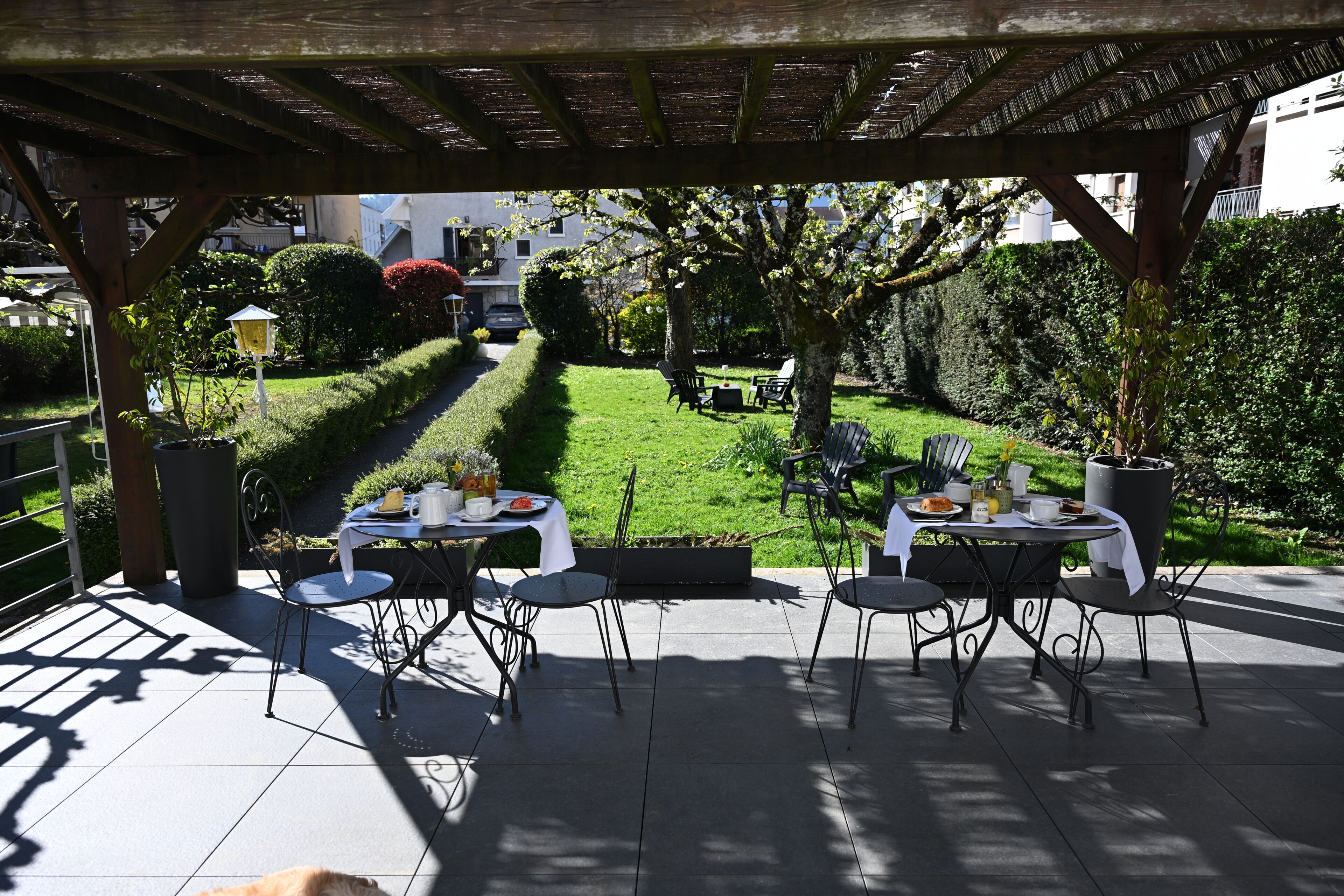 An outdoor patio with tables and chairs, a pergola overhead, and a well-maintained garden in the background.