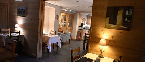 A restaurant with wooden walls, tables with white tablecloths, and a kitchen area visible through an open doorway.