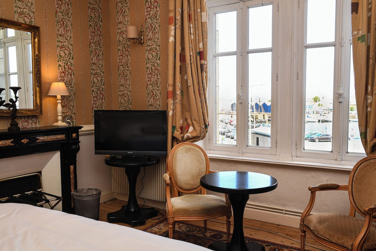 A hotel room with a bed, a TV, a chair, a small table, and a view of a marina through large windows.