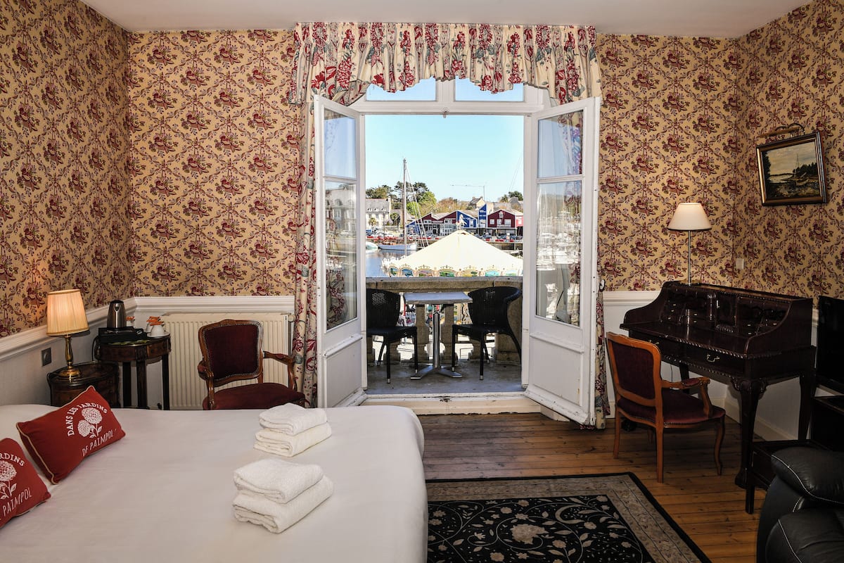 A hotel room with a bed, a piano, a table with chairs, and a view of the harbor through an open door.