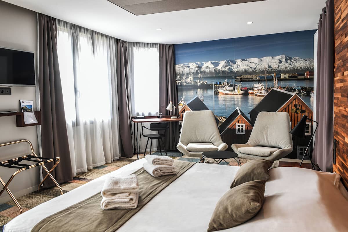 A hotel room with a bed, two armchairs, a desk, and a large window with a scenic view of a harbor and mountains.