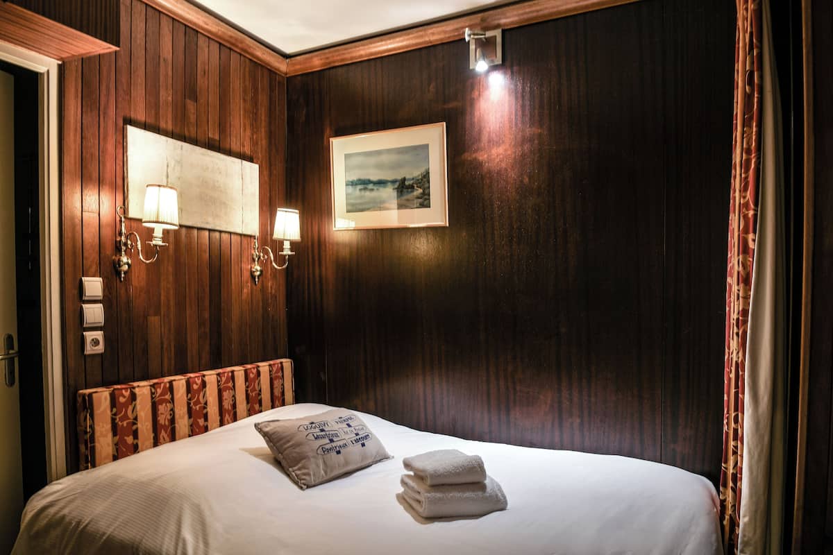 A hotel room with a wooden headboard, a bed with white linens, a pillow, a folded towel, and a framed picture on the wall.