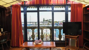 A room with a large window, a wooden table with a vase, a television on a wooden stand, and a chair. - Le K'Loys (Paimpol)