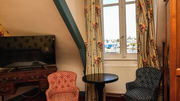 A room with a TV on a wooden dresser, two patterned chairs, a small round table, and a window with curtains.