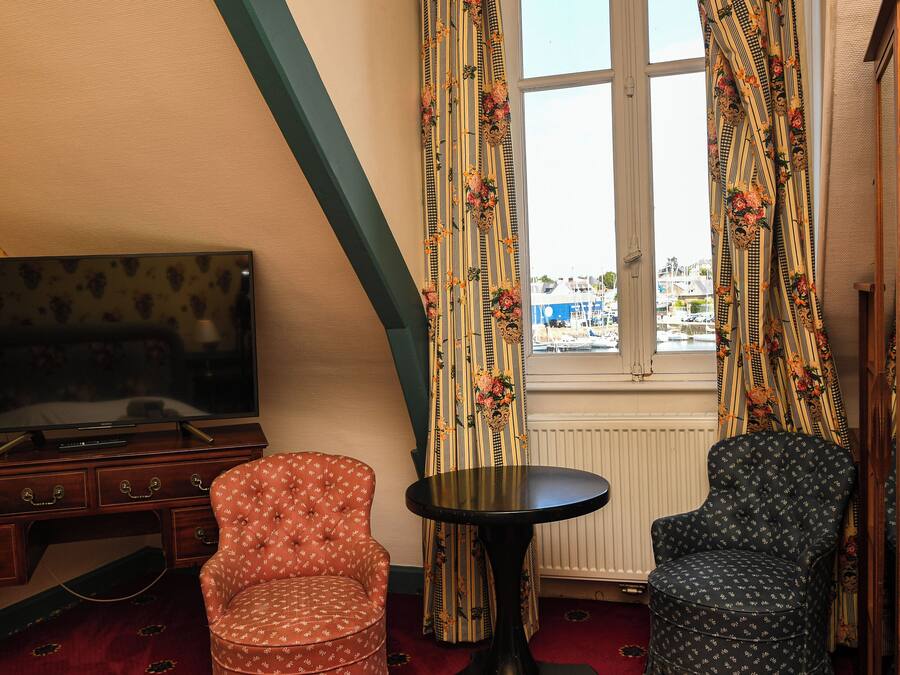 A room with a TV on a wooden dresser, two patterned chairs, a small round table, and a window with curtains.